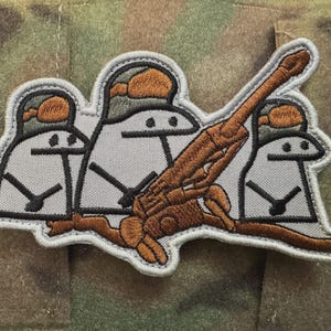 May include: Embroidered patch showing three cartoon figures in helmets operating a brown artillery cannon. The figures are white with black details, and the patch has a grey border. The background is camouflage fabric.