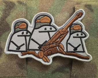 May include: Embroidered patch featuring three cartoon figures with helmets operating a brown artillery cannon. The figures are white with black details, and the patch has a gray border. The background is camouflage fabric.