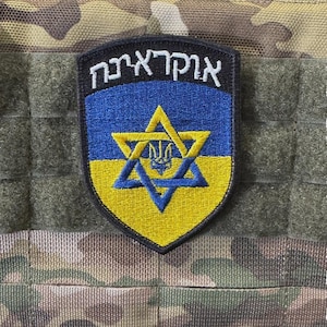 Puede incluir: Parche en forma de escudo con la palabra hebrea "אוקראינה" en blanco sobre una estrella de David azul y amarilla. El parche está sobre un fondo de camuflaje.