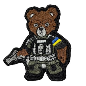 May include: Embroidered patch of a brown teddy bear in military attire, including a tactical vest and camouflage trousers. The bear holds a silver pistol and has a blue and yellow armband. The patch has a black border.