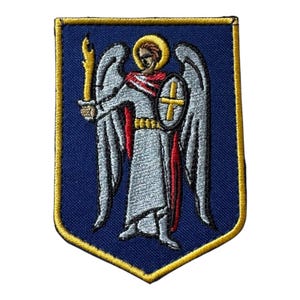 May include: A shield-shaped embroidered patch featuring a dark blue background with a gold border. The design depicts a winged figure in a light grey robe with a red sash, holding a yellow torch in one hand and a shield with a gold cross.