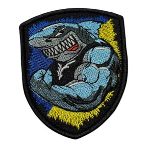 May include: Embroidered patch depicting a strong shark with a menacing grin. The shark is in blue and grey, wearing a black top. The background has blue and yellow accents, within a black border.