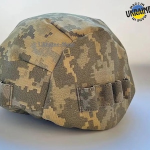 May include: A tactical helmet cover featuring a digital camouflage pattern in shades of tan, brown, and olive green. The cover includes integrated fabric loops along the sides and a circular logo in the upper right corner with the text Made in Ukraine.