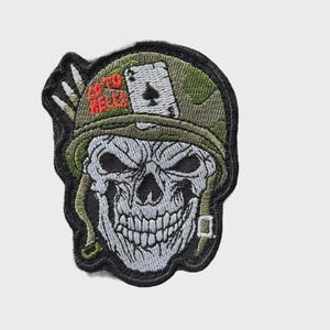 Puede incluir: Parche bordado con una calavera que lleva un casco de camuflaje, con una carta de juego y el texto "GO TO HELL". La calavera es blanca con sombreado detallado, y el parche tiene un borde negro.