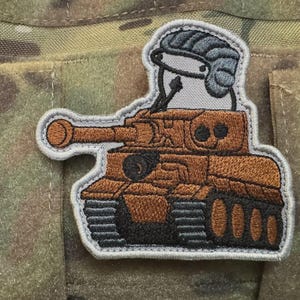 May include: Embroidered patch featuring a cartoon character in a tank. The tank is brown with black details and a long barrel. The character wears a gray helmet. The patch has a gray border and is set against a camouflage background.