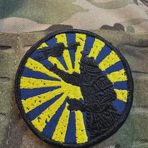 May include: A round patch featuring a black samurai warrior silhouette against a yellow sunburst design on a blue background. The patch is embroidered with a textured appearance and is set against a camouflage background.