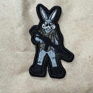 Può includere: Toppa ricamata raffigurante un coniglio dei cartoni animati in tenuta militare, con un fucile. La toppa ha un bordo nero, con il coniglio in sfumature di grigio, verde e beige. La toppa è alta circa 7,6 cm.
