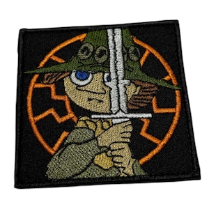 May include: Embroidered patch featuring a cartoon character with a sword, wearing a green hat and olive-green clothing. The character is set against a black background with an orange, stylized sun or starburst design.