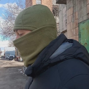 May include: An olive green fleece balaclava that covers the head and neck, featuring a fitted design with a single opening for the eyes and nose. The fabric has a soft, matte texture and is worn with a black hooded jacket with a grey inner lining.