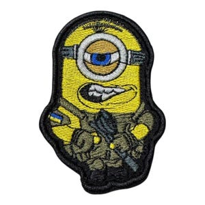 Puede incluir: Parche bordado con un personaje Minion amarillo con un solo ojo azul, vestido con uniforme militar y sosteniendo un rifle. El parche tiene un borde negro e incluye una pequeña bandera ucraniana. El Minion tiene una expresión enfadada.
