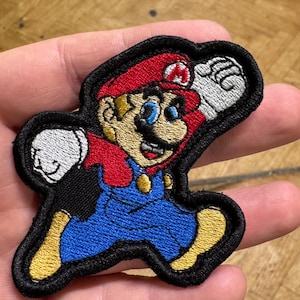 Peut inclure: Écusson brodé représentant le personnage de Mario de la série de jeux vidéo Super Mario Bros. Mario est représenté dans sa tenue classique : un chapeau rouge, une salopette bleue et des gants blancs, avec une bordure noire.