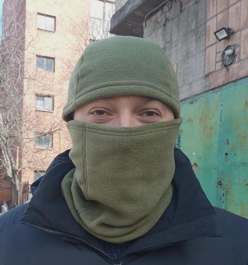 May include: An olive green fleece balaclava that covers the head and neck, featuring a rounded cap section and a face mask portion that leaves the eyes exposed. The fabric has a soft, textured finish with visible stitching along the seams.
