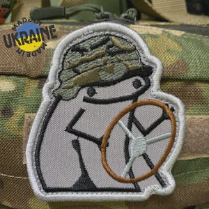 Könnte beinhalten: Gestickter Aufnäher mit einer Cartoonfigur mit Tarnhut und Lenkrad. Der Aufnäher hat einen grauen Hintergrund mit schwarzen und braunen Details und einen weißen Rand. Der Aufnäher befindet sich auf einem Tarnhintergrund.