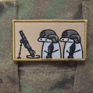 Puede incluir: Parche bordado con un mortero de dibujos animados y dos figuras con cascos. El parche tiene un fondo beige con un borde marrón, sobre una tela de camuflaje. El texto "MADE IN UKRAINE" es visible.
