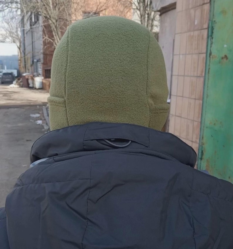 May include: An olive green fleece beanie with a multi-panel construction and stitched seams, worn above a black jacket with a high collar. The hat features a soft, textured fabric surface that covers the head and ears, providing a snug fit.