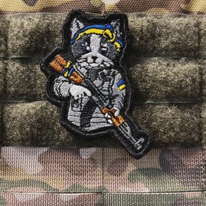 May include: Embroidered patch featuring a cat in military attire, holding a rifle. The cat is gray and white, wearing a blue and yellow headband. The patch has a black border and is set against a camouflage background.
