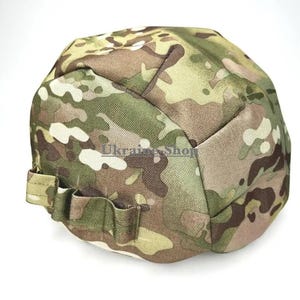 May include: A camouflage helmet cover featuring a multi-colored pattern of green, brown, and tan shapes. The fabric cover includes a series of stitched loops along the side and displays the text "MADE IN UKRAINE" in a circular logo at the top.