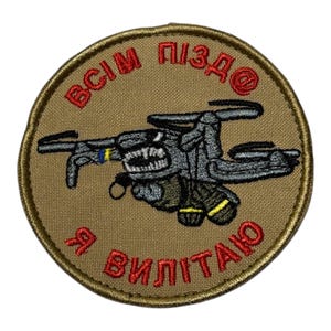 May include: Embroidered patch featuring a stylized drone with a menacing expression. The patch has text in red, and the drone has a yellow and blue accent. The patch is circular with a tan background and a brown border.