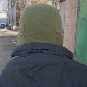 May include: An olive green fleece beanie with a multi-panel construction and stitched seams, worn above a black jacket with a high collar. The hat features a soft, textured fabric surface that covers the head and ears, providing a snug fit.