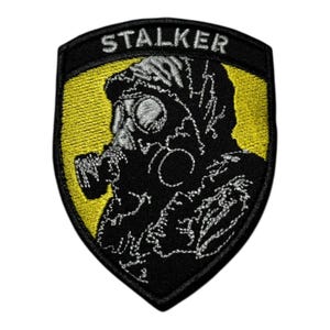 Può includere: Toppa ricamata con una silhouette nera di una persona con maschera antigas su sfondo giallo. La parola "STALKER" è ricamata in bianco sopra la figura. La toppa ha un bordo nero.