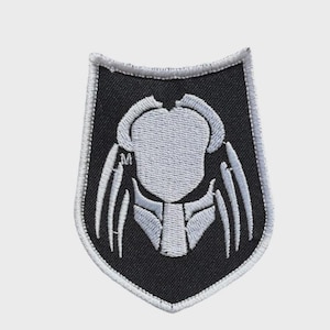 Pode incluir: Um patch bordado em preto e branco com o design da máscara do Predator. O patch tem formato de escudo com uma borda branca. A máscara do Predator é branca, com linhas e formas detalhadas, sobre um fundo preto.