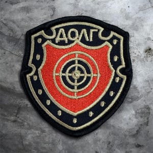 May include: A shield-shaped embroidered patch featuring a black border with light-colored dots, a bright orange center, and a central target graphic. The Cyrillic text "ДОЛГ" is stitched in light-colored thread at the top of the black section.