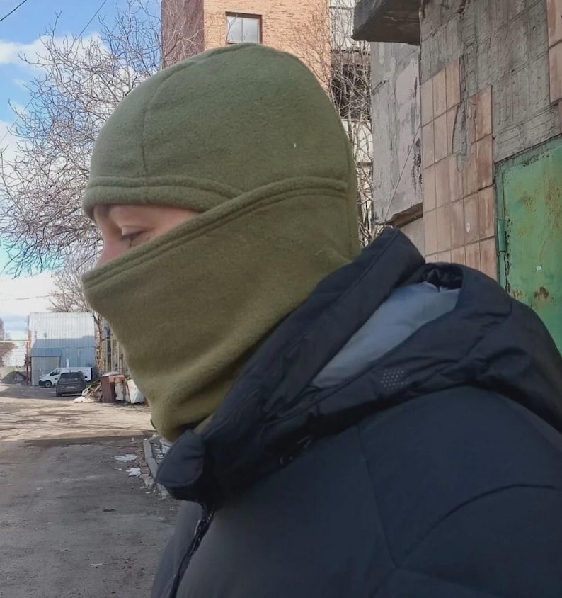 May include: An olive green fleece balaclava that covers the head and neck, featuring a fitted design with a single opening for the eyes and nose. The fabric has a soft, matte texture and is worn with a black hooded jacket with a grey inner lining.