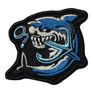 Puede incluir: Parche bordado que representa un tiburón mordiendo un ancla, con un diseño en tonos azules, blancos y negros. El parche tiene un borde negro y es adecuado para ropa o accesorios.