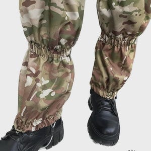 Puede incluir: Un par de polainas de camuflaje con un patrón multicolor de verde, marrón y beige. Las polainas tienen una sección central elástica y un cierre de cordón ajustable en la parte superior, usadas sobre botas negras con suelas de goma gruesas.