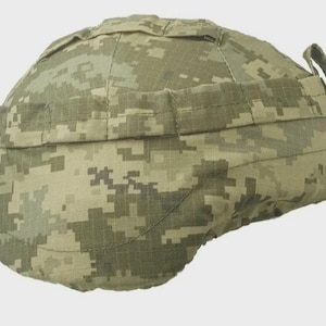 May include: A tactical combat helmet cover featuring a digital camouflage pattern in shades of olive green, tan, and brown. The cover includes integrated fabric webbing straps across the exterior surface for attaching gear and accessories.