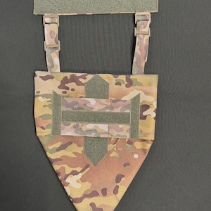 Peut inclure: Accessoire tactique à motif camouflage avec sangles réglables et fermeture auto-agrippante. L'accessoire présente un fond triangulaire et un dessus rectangulaire avec une bande auto-agrippante verte. Les sangles ont des boucles en plastique.