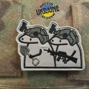 Può includere: Toppa ricamata con due personaggi dei cartoni animati con elmetti militari, uno con una chiave inglese e l'altro con un fucile. La toppa è grigia e bianca con dettagli neri, e il testo "MADE IN UKRAINE" è sopra.