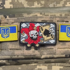 May include: A tactical vest with three patches. Two patches feature the Ukrainian flag in blue and yellow. The central patch depicts a skull-faced jester holding a playing card with a spade.
