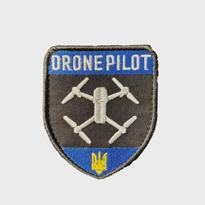May include: A shield-shaped patch with the text "DRONE PILOT" in white on a blue background. The patch features a white drone graphic and a yellow trident symbol on a blue base. The patch has a black border.