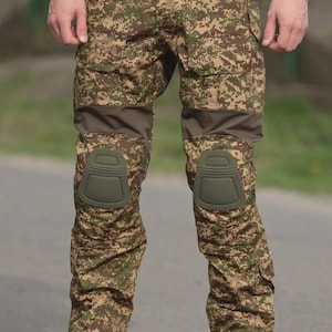 Pode incluir: Calças cargo camufladas com joelheiras. As calças apresentam um padrão pixelizado em tons de castanho, verde e bege. As joelheiras são verde-oliva. As calças têm vários bolsos e são combinadas com botas bege.