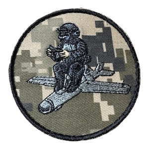 Op de afbeelding: Een ronde geborduurde patch met een zwarte beer in een helm en vlieguitrusting, zittend op een grijs vliegtuig. De achtergrond bestaat uit een beige, bruin en donkergroen digitaal camouflagepatroon met een dikke zwarte rand.