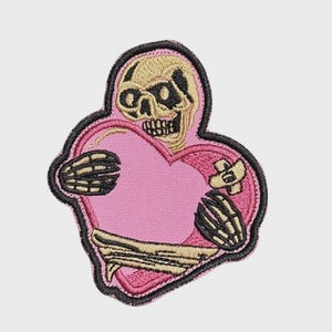 Può includere: Toppa ricamata con un teschio che abbraccia un cuore rosa. Il teschio e le mani scheletriche sono beige, con un contorno nero. Il cuore ha una benda e un bordo nero. Un design unico e stravagante.