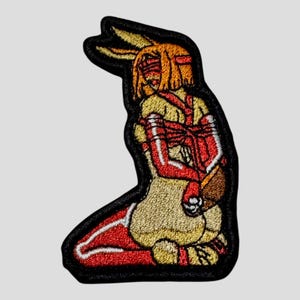 Puede incluir: Parche bordado que representa una figura arrodillada con orejas de conejo, en tonos amarillos, rojos y marrones. La figura está atada con correas rojas, delineada en negro y tiene un borde negro.