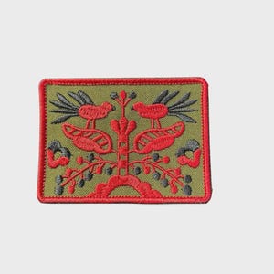 May include: Embroidered patch with a rectangular shape. The patch features a design of two red birds perched on a stylized tree with red and gray leaves and branches. The background is olive green, and the border is red.