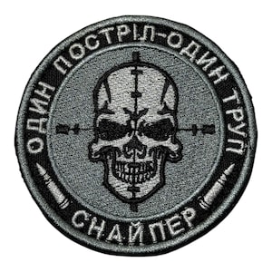 Könnte beinhalten: Ein runder, grauer Aufnäher mit schwarzem, gesticktem Text und einem Totenkopf-Design. Der Totenkopf hat ein Fadenkreuz. Der Text lautet "ОДИН ПОСТРІЛ - ОДИН ТРУП СНАЙПЕР" und ist kreisförmig um den Totenkopf angeordnet.
