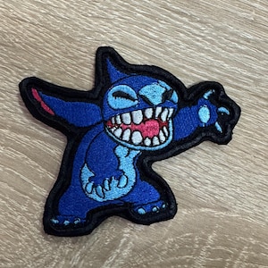 Peut inclure: Un écusson brodé représentant un personnage extraterrestre bleu avec de grandes oreilles, un large sourire montrant des dents blanches pointues et une langue rose. Le personnage est dans une pose dynamique, bordé de fil noir épais.