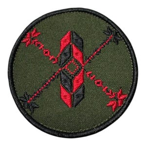 Peut inclure: Écusson brodé de forme circulaire. Le patch présente un motif central en forme de plume noire et rouge, croisé par deux flèches rouges aux extrémités décoratives. Le fond est vert olive et le patch a une bordure noire.