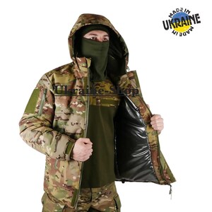 Op de afbeelding: Een tactisch camouflagejack met capuchon, voorzien van een donker olijfgroene nekwarmer en een zwarte binnenvoering. Het jack heeft een ritssluiting en een klittenbandvlak op de mouw. Een cirkelvormig logo toont de tekst Made in Ukraine.
