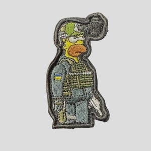 May include: Embroidered patch featuring a cartoon character in military gear. The character has yellow skin, an orange beard, and is wearing a green helmet and vest. The patch includes the Ukrainian flag and is outlined in gray.