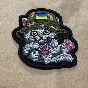 May include: Embroidered patch of a grey cat wearing a camouflage bucket hat with a Ukrainian flag. The cat has pink paw pads and is outlined in black.
