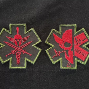 May include: Two embroidered patches with a black background. One patch features a red Spartan helmet, crossed swords, and a caduceus. The other patch has a red skull, a heartbeat line, and the word "MEDIC". Both patches have a green border.