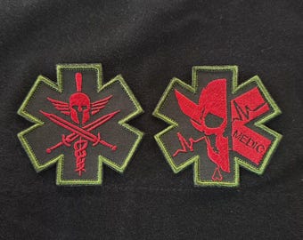 May include: Two embroidered patches with a black background. One patch features a red Spartan helmet, crossed swords, and a caduceus. The other patch has a red skull, a heartbeat line, and the word "MEDIC". Both patches have a green border.