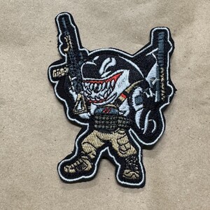 Könnte beinhalten: Gestickter Aufnäher mit einem Cartoon-Orca mit bedrohlichem Grinsen, der zwei Gewehre hält. Der Orca trägt taktische Ausrüstung und eine khakifarbene Hose. Der Aufnäher hat einen schwarzen Rand und befindet sich auf einem beigen Hintergrund.