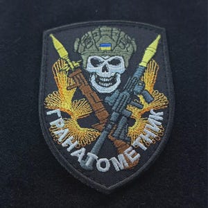 Puede incluir: Parche bordado negro con una calavera con casco y bandera ucraniana. Incluye un rifle, un lanzacohetes y texto cirílico. Detalles dorados, marrones, azules y blancos.