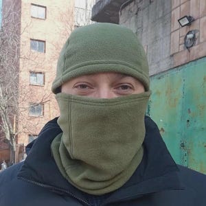 May include: An olive green fleece balaclava that covers the head and neck, featuring a rounded cap section and a face mask portion that leaves the eyes exposed. The fabric has a soft, textured finish with visible stitching along the seams.